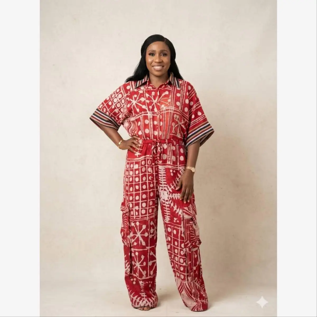 A woman stands confidently in a full-length handcrafted adire batik jumpsuit featuring a vivid crimson red and off-white resist-dye pattern with geometric motifs, polka dots, and starburst designs. The outfit includes a wide-leg cargo silhouette, a drawstring waist, and striped cuff detailing on the short sleeves. Color: Crimson Red and Off-White