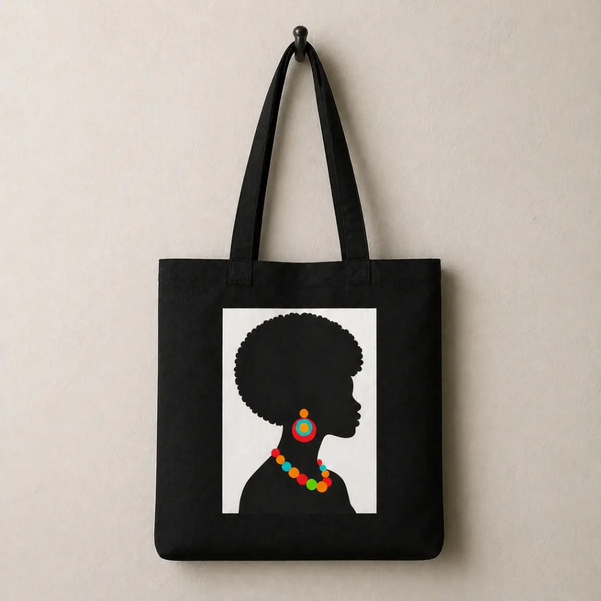 Front flat-lay view of a black canvas tote bag hanging on a wall hook, featuring a bold white-paneled print of an African woman silhouette with a natural afro, colorful hoop earring, and multicolor beaded necklace in red, orange, green, and teal. The bag has black fabric handles and a clean, structured shape. Color: Black