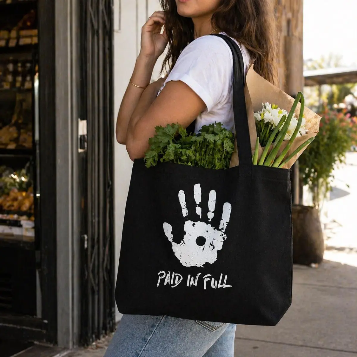 A woman carries a large black canvas tote bag over her shoulder while walking past a market storefront, with fresh herbs and flowers visible inside the bag. The bag features a bold white distressed handprint graphic with the words 'Paid In Full' printed below it. Color: Black