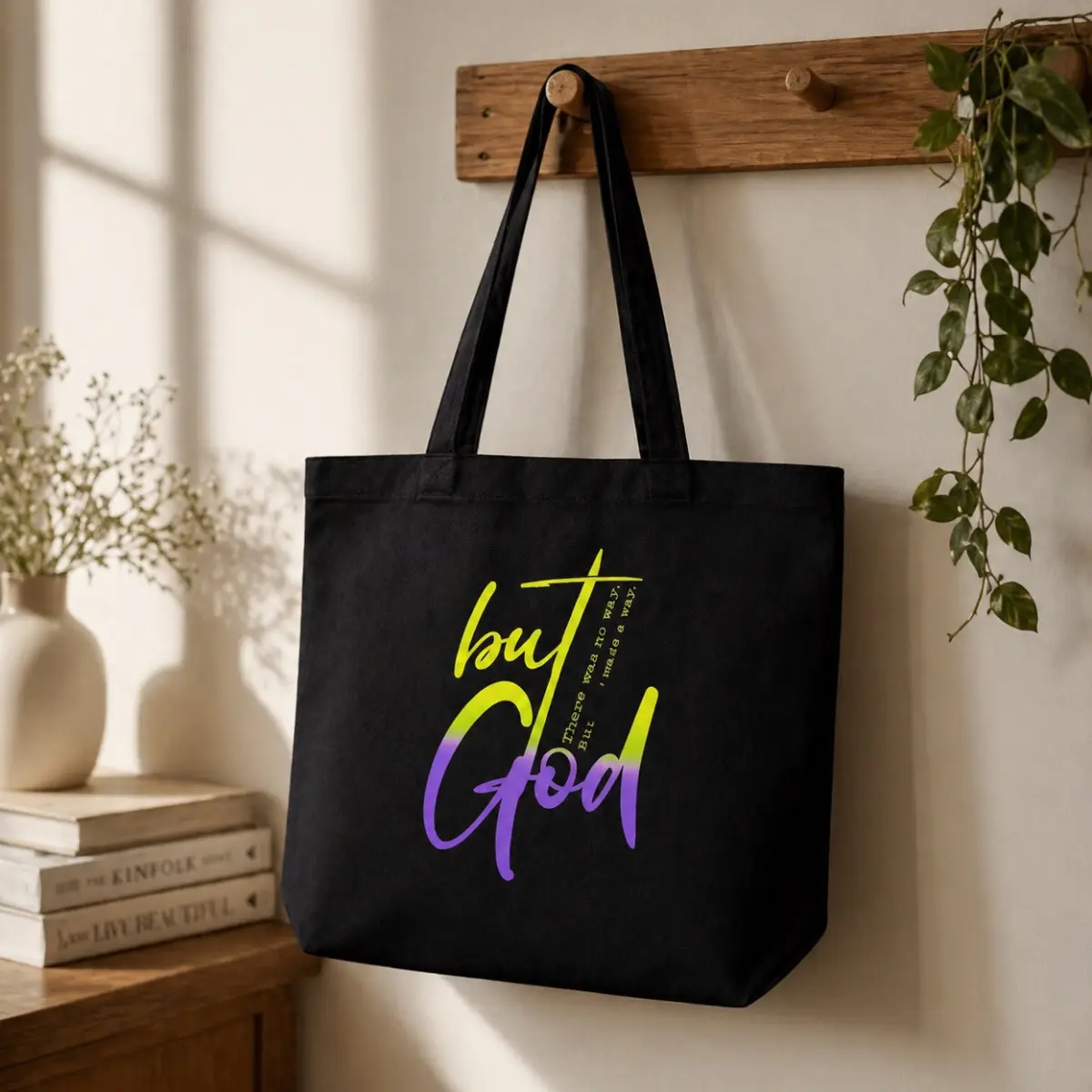 A black canvas tote bag hangs from a wooden wall hook, featuring a bold hand-styled graphic print that reads 'but God' in expressive yellow-green and purple brush lettering, with the inspirational subtitle 'There was no way, But God made a way.' The artisan-designed print showcases vibrant color gradients and dynamic typography on sturdy, quality canvas fabric with long black shoulder straps. Styled in a warm home setting with books and trailing greenery, this faith-inspired tote is ideal for everyday carry with a meaningful message. Color: Black, Yellow-Green, Purple