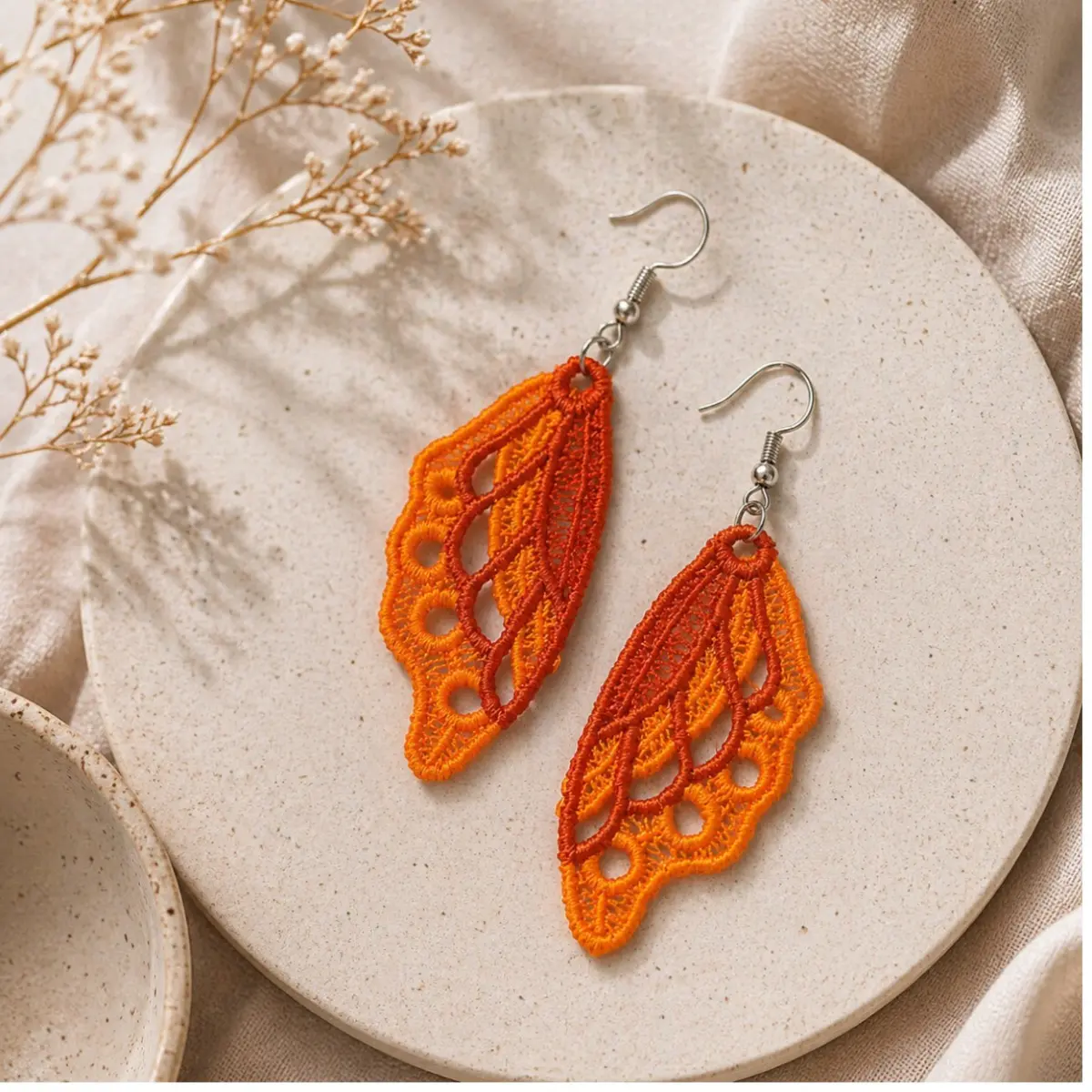 A pair of handcrafted butterfly wing dangle earrings displayed on a speckled ceramic tray with dried floral stems in the background. The earrings feature intricate lace-like embroidered threadwork in a delicate open cutwork pattern, showcasing skilled artisan craftsmanship with layered light and dark orange threads. Silver-toned fish hook ear wires complement the lightweight textile design, measuring approximately 5 cm long by 2.5 cm wide. Color: Light Orange, Dark Orange
