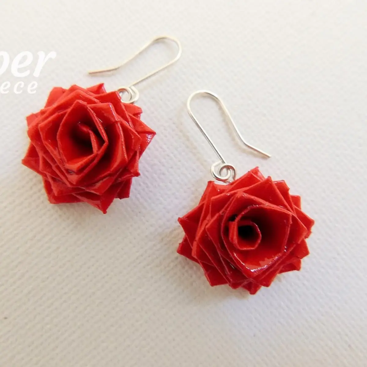 A pair of artisan-made crimson red origami rose drop earrings photographed side by side on a light linen surface, highlighting the tight spiral center and sharp angular petal edges of each hand-folded paper rose bloom on silver earring hooks. Color: Red